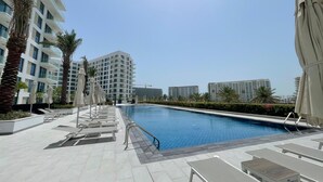 A swimming pool area with lounge chairs and umbrellas, surrounded by modern buildings. - Stylish Studio Apartment with Kitchen, TV Lounge & Dining Space (Muḥāfaẓat al-Muḥarraq)