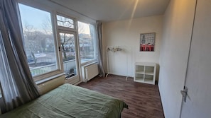 A room with a bed, a radiator, a window with curtains, a coat rack, and a small white shelf.