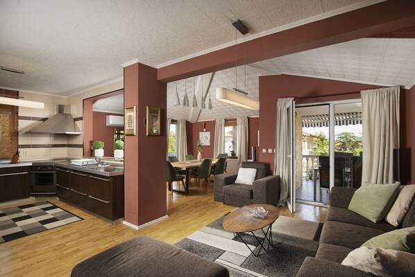 Eine moderne Küche mit dunklen Schränken, ein Essbereich mit Tisch und Stühlen, ein Wohnzimmer mit Sofa und Couchtisch sowie eine Glastür, die auf eine Terrasse im Freien führt.