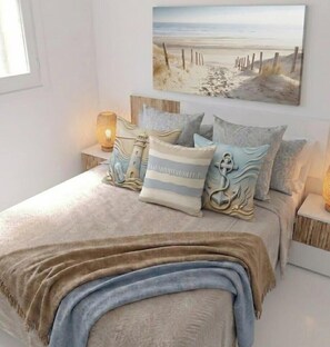 Ein Schlafzimmer mit einem Bett, Kissen, einer Decke, einem Strandbild und Nachttischlampen.