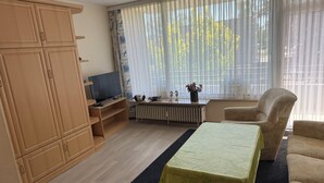 Ett vardagsrum med en TV, träskåp, ett element, en soffa och ett fönster med tunna, genomskinliga gardiner.