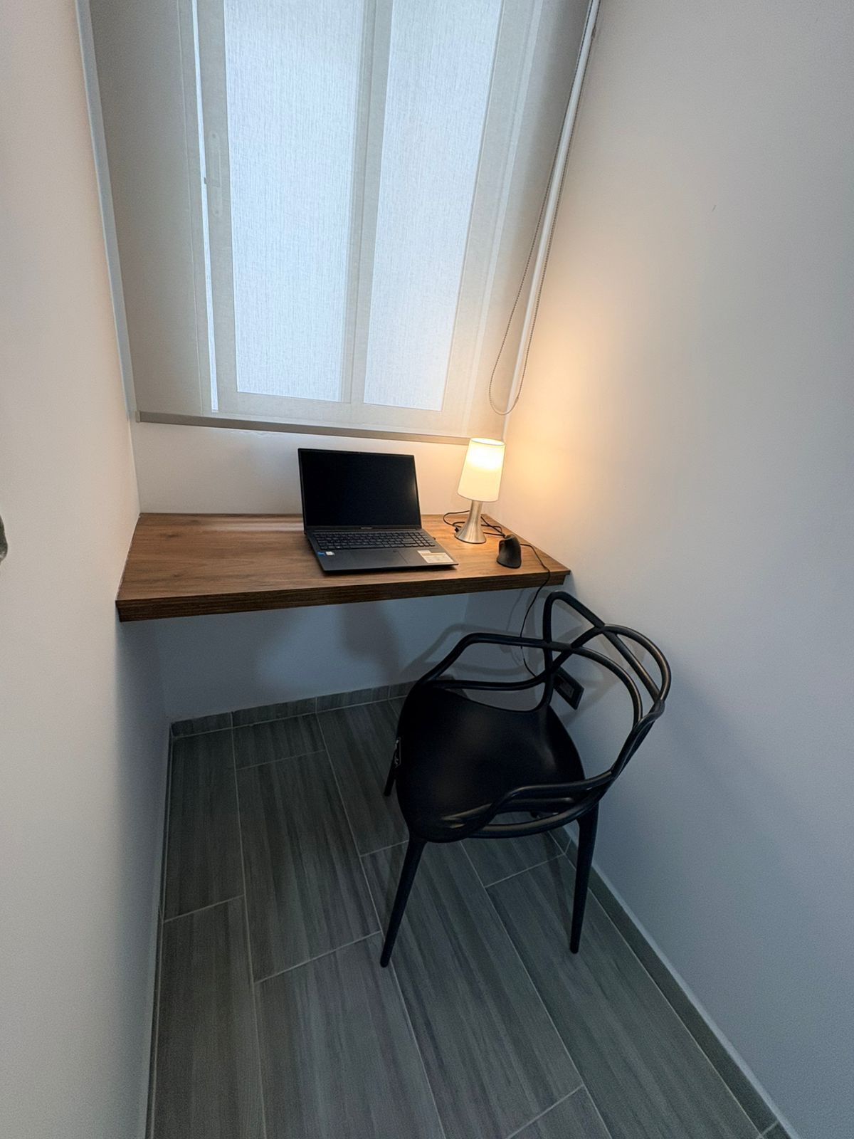 A small workspace with a laptop on a wooden shelf, a desk lamp, and a black chair.