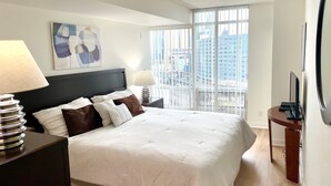 Ein Hotelzimmer mit einem großen Bett, zwei Nachttischen mit Lampen, einem Fernseher und Blick auf die Stadt durch raumhohe Fenster.