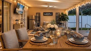 Un área de comedor al aire libre con una mesa de madera dispuesta para cuatro personas, rodeada de sillas, una televisión montada en la pared y un cartel que dice 'happy place'.