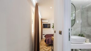 Moderni hotellihuone, jossa on kalanruotokuviollinen lattia, ruskealla peitteellä varustettu sänky, seinälle asennettu televisio ja lasinen suihkukaappi.