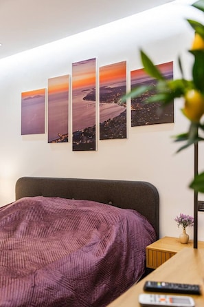 Hotelkamer met een bed, nachtkastje, afstandsbediening en een wandtelevisie die uitzicht geeft op een zonsondergang.