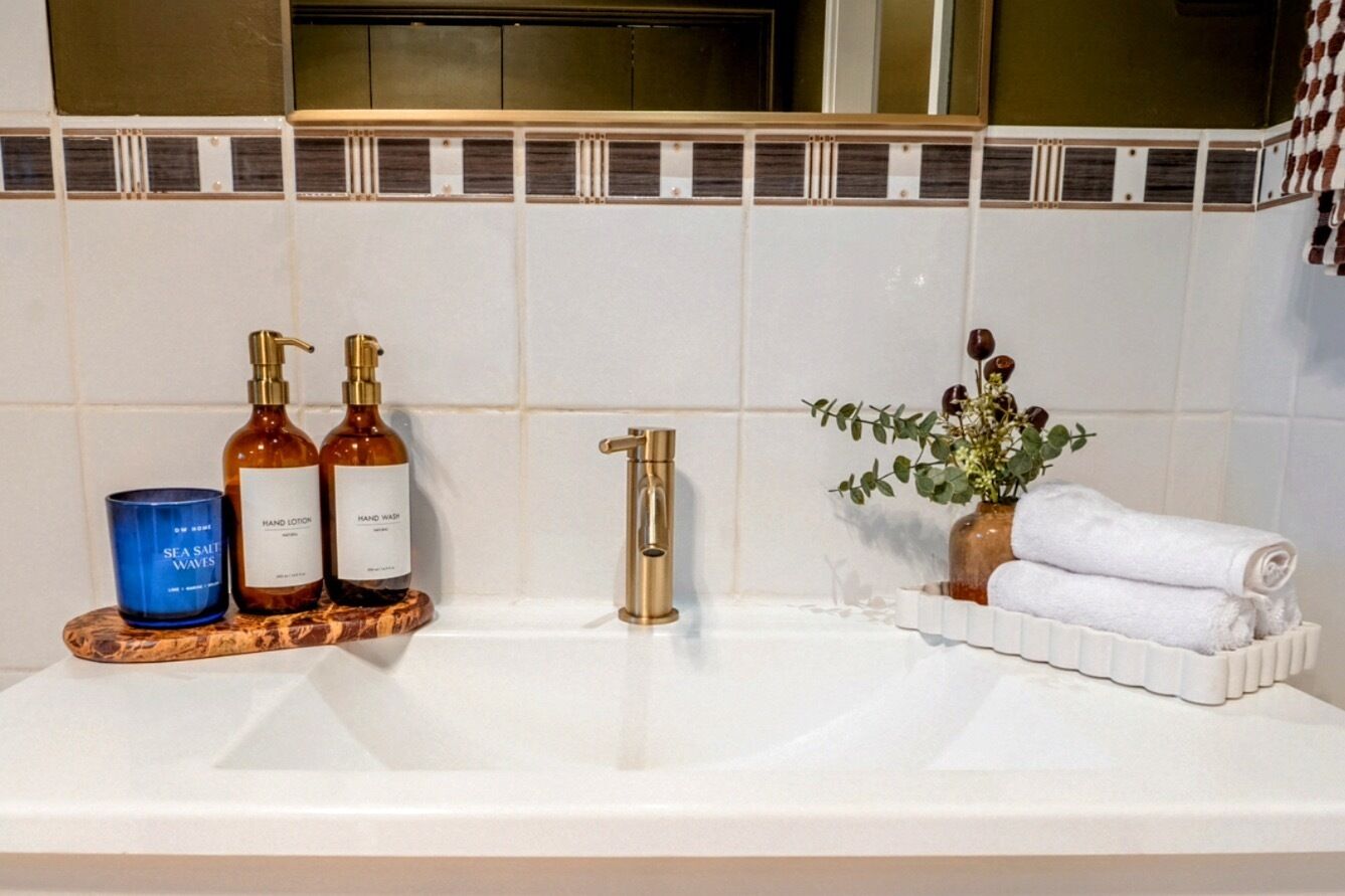 Un tocador de baño con encimera blanca, grifo dorado y dos botellas de crema para manos.