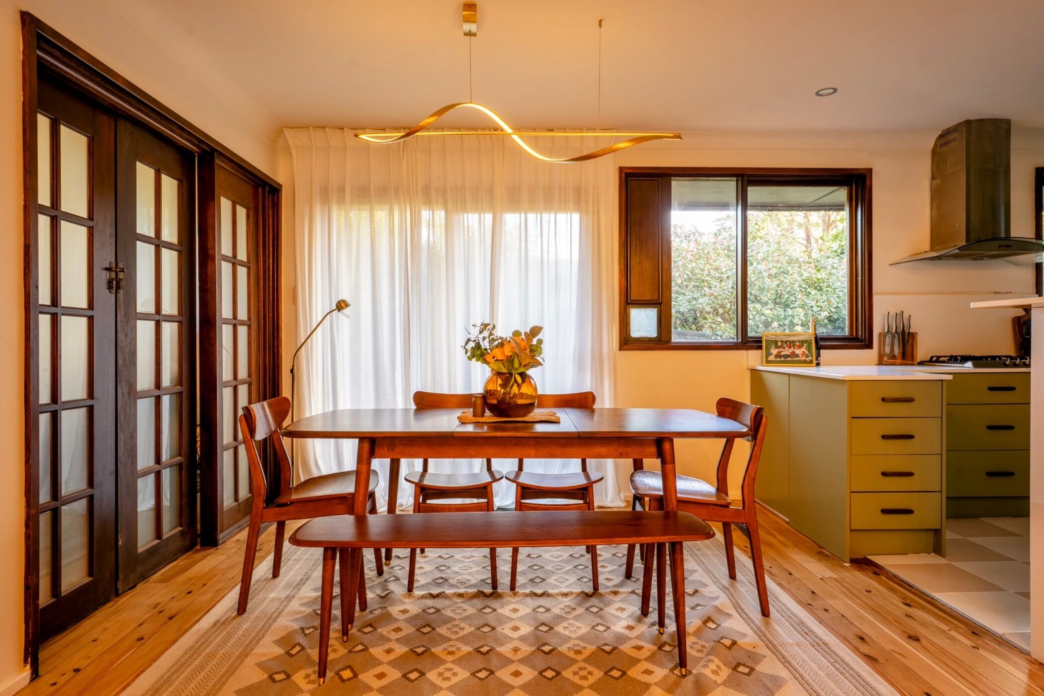 Un comedor con una mesa y sillas de madera, un florero con flores, y una ventana con cortinas.