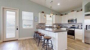 A kitchen with white cabinets, a central island with stools, stainless steel appliances, and a window with blinds. - Candy's Cove: 2  BR, 1  BA House in Crystal Beach, Sleeps 8 (Crystal Beach)