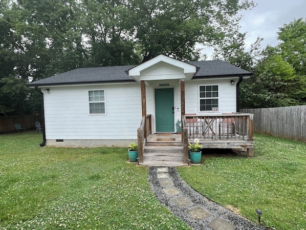 A small white house with a green door, a porch, and a fenced backyard.