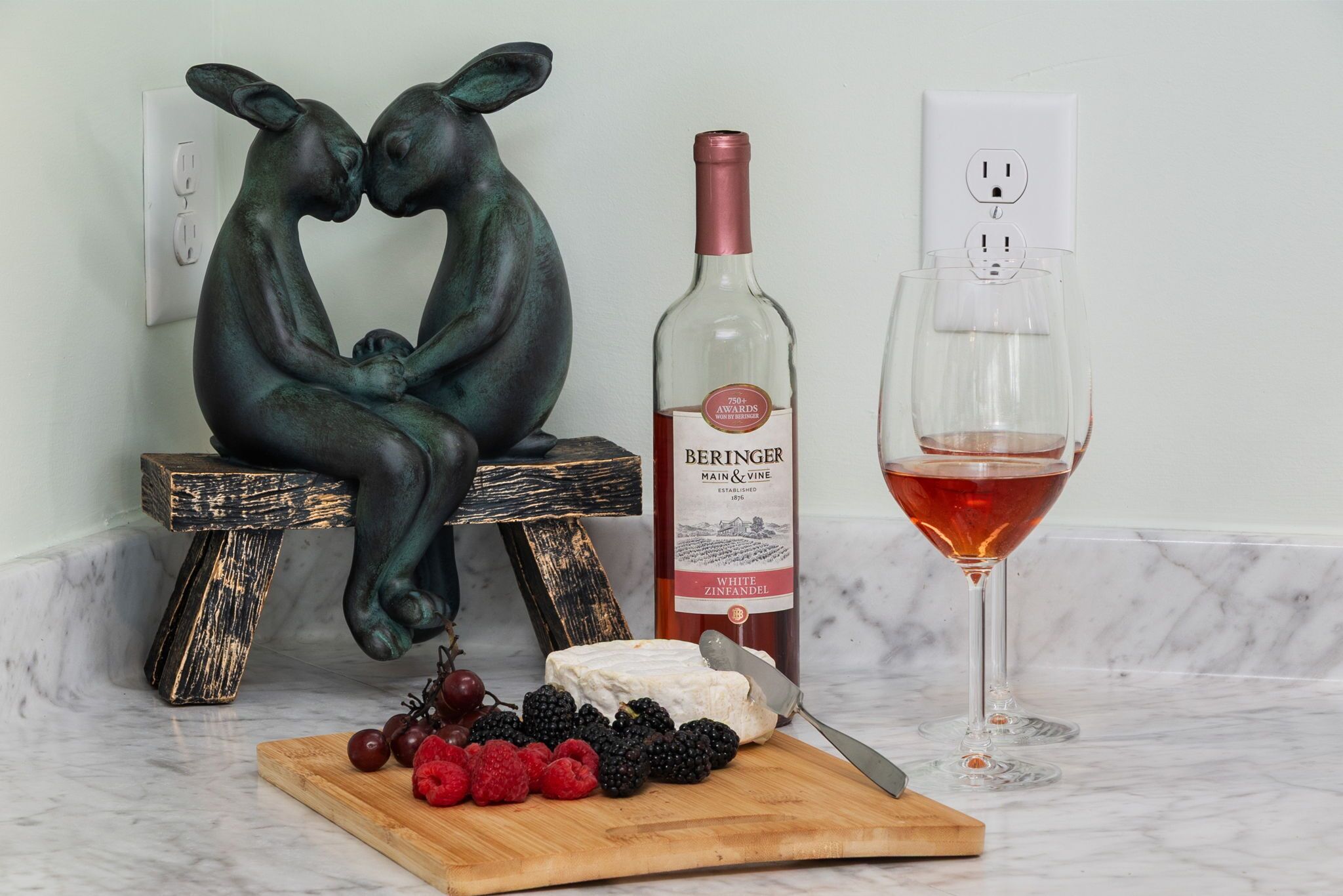 En bordplade i marmor med en flaske Beringer vin, et glas rødvin, en skærebræt med frugt og en skulptur af to figurer.