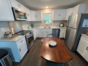 A compact kitchen with white cabinets, stainless steel appliances, a wooden dining table, and a window with a view of greenery.