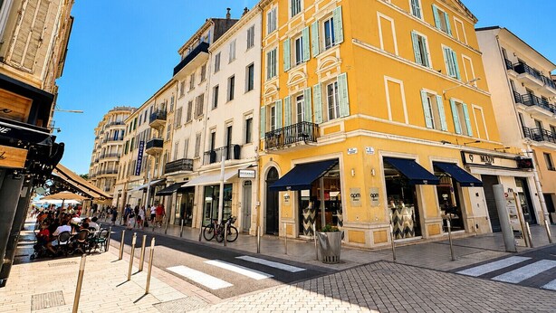 Un immeuble à plusieurs étages, doté d’une façade jaune, de boutiques au rez-de-chaussée et d’un espace piétonnier avec des places assises en plein air.