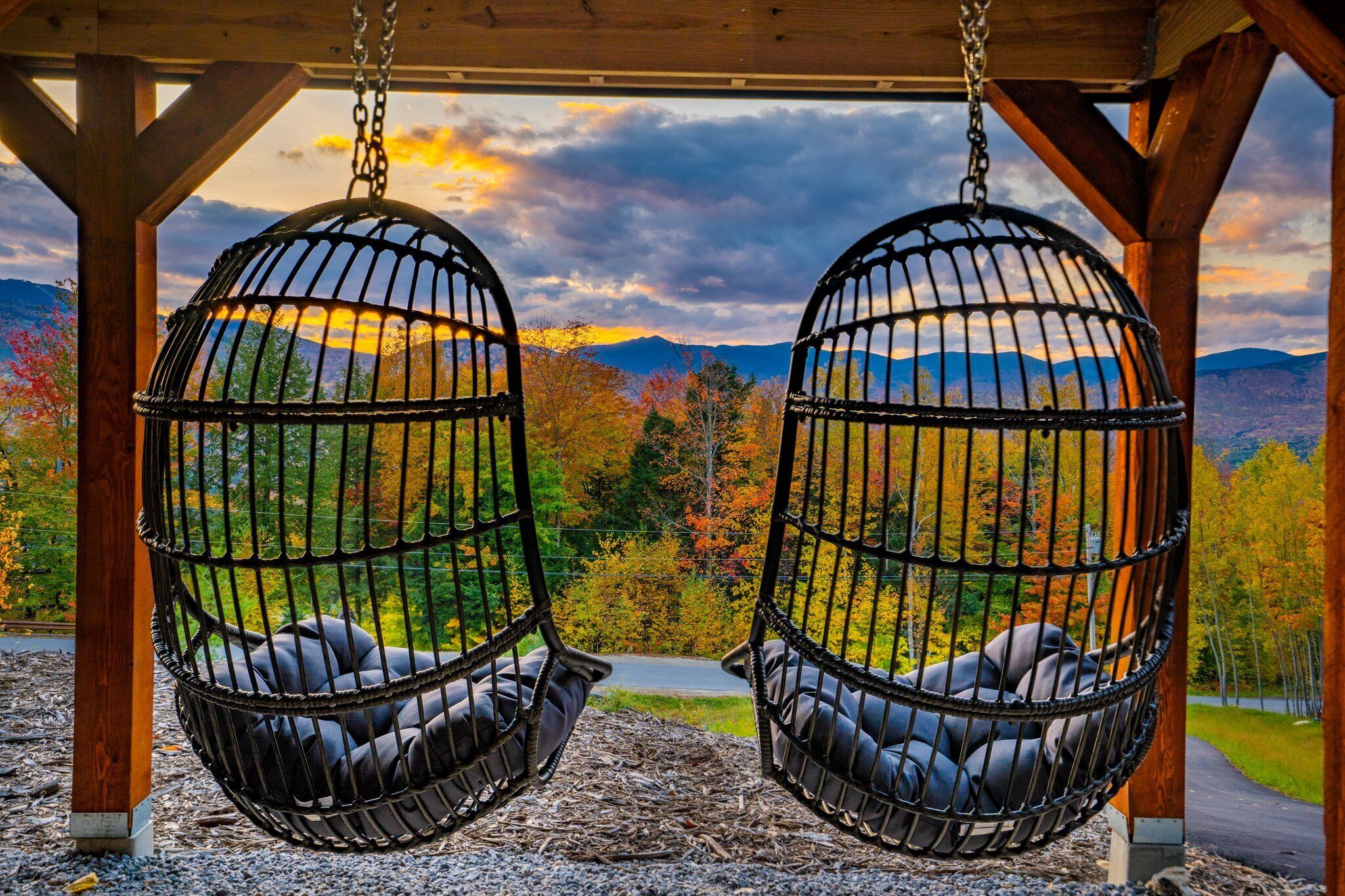 Two black swing chairs with cushions, suspended from a wooden structure with a view of autumn foliage and mountains.. Spacious 5-bedroom chalet with slope views of Sunday River slopes