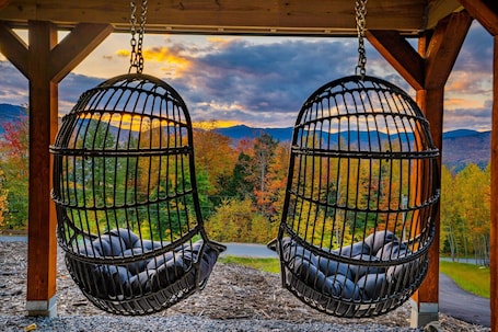 Two black swing chairs with cushions, suspended from a wooden structure with a view of autumn foliage and mountains.. Spacious 5-bedroom chalet with slope views of Sunday River slopes