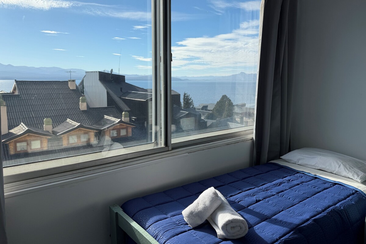 Una cama con una colcha azul y una toalla enrollada, una ventana con vistas a edificios y montañas, y una cortina entreabierta.