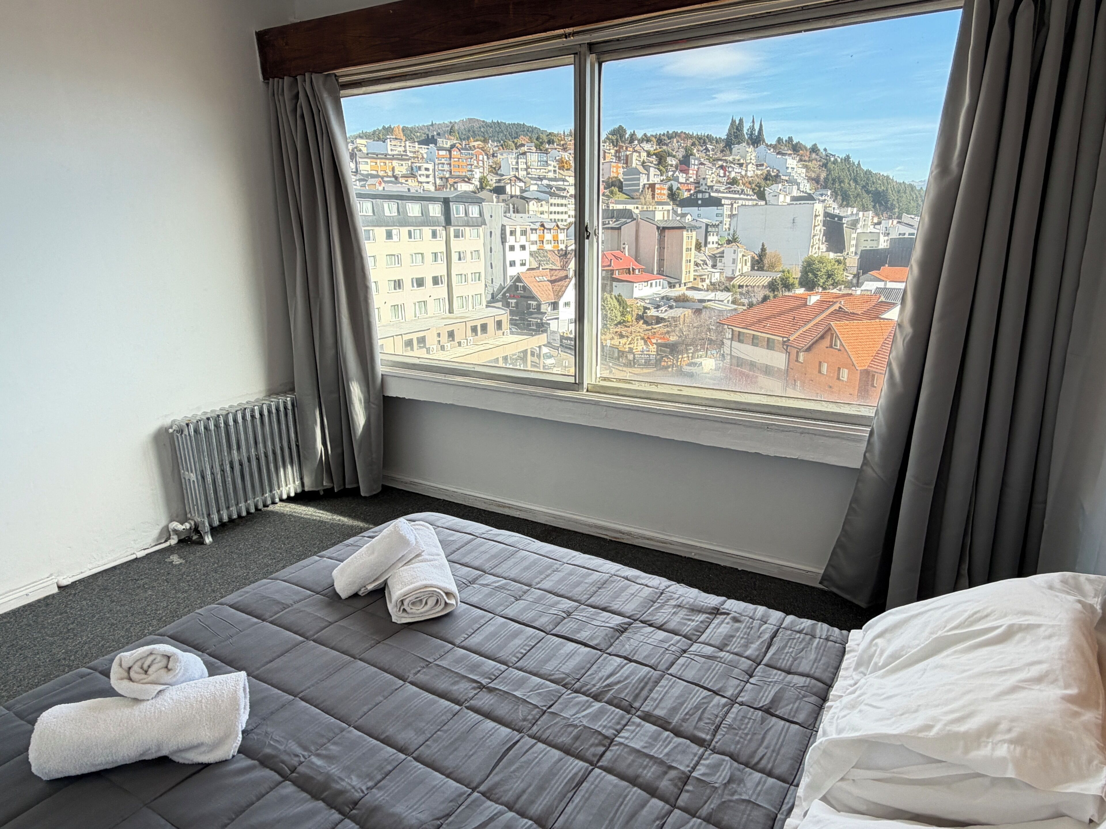 A hotel room with a large window, a bed with a grey quilt, and two towels on the bed.