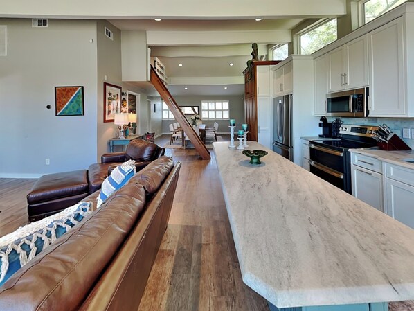 A spacious kitchen with a long island, modern appliances, and a staircase leading to an upper level. - Serenity View | Fenced Yard, Private Dock, Pet-Friendly (Tybee Island)