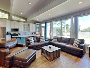 A spacious living area with a leather sectional sofa, ottomans, and a coffee table. A kitchen with white cabinets and a microwave is visible in the background. - Serenity View | Fenced Yard, Private Dock, Pet-Friendly (Tybee Island)