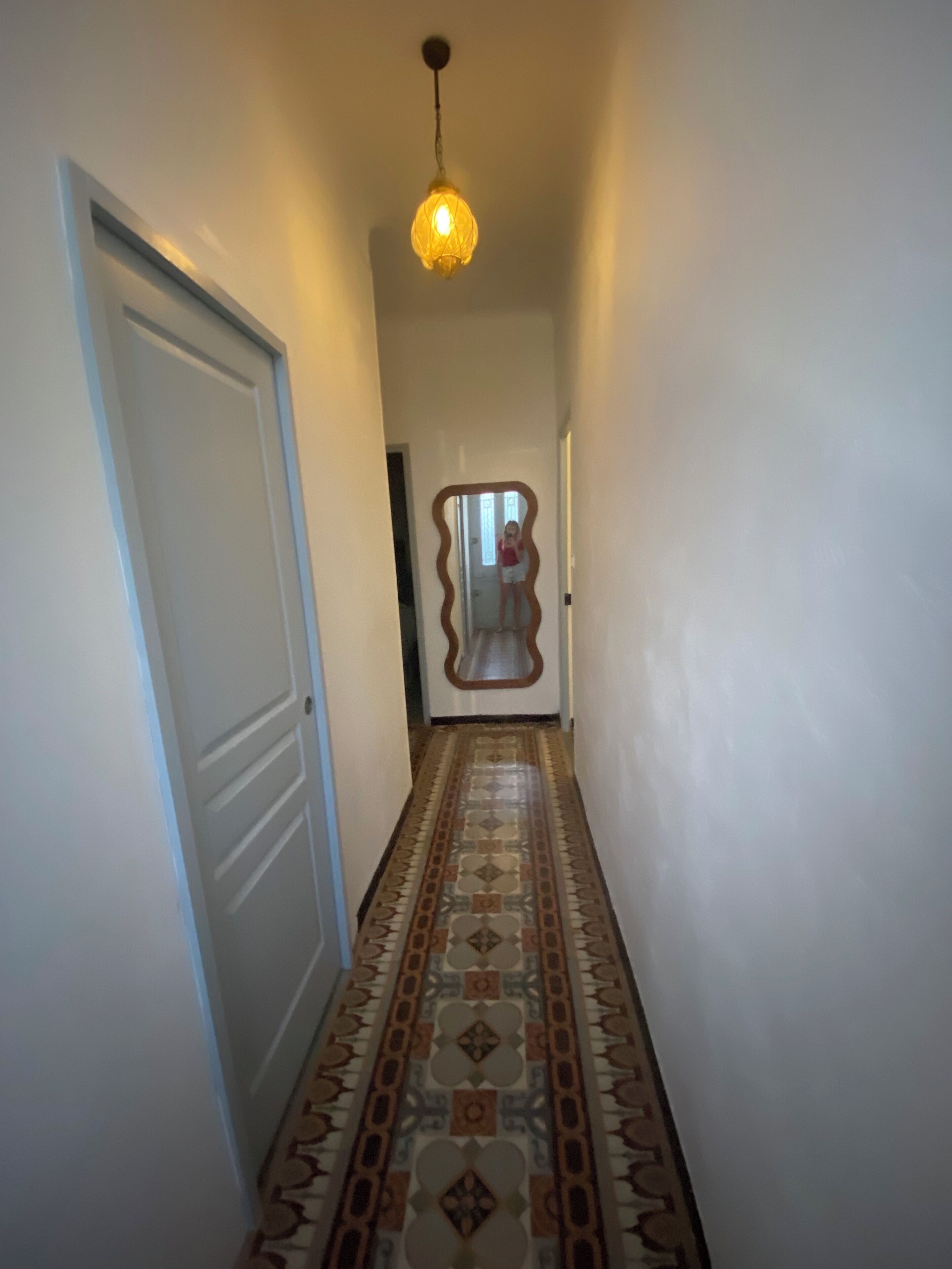 Un couloir avec un sol à motifs, une porte sur la gauche et un luminaire suspendu au plafond.