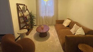Uma sala de estar com um sofá marrom, uma mesa de centro redonda, uma mesinha de madeira, uma estante e um lustre.