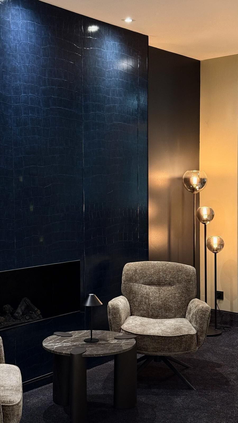 Un interior moderno con una pared texturizada oscura, una mesa redonda con una lámpara, un sillón mullido y lámparas de pie.