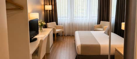 Habitación de hotel con cama, escritorio con televisor, dos sillas, espejo y ventana con cortinas.