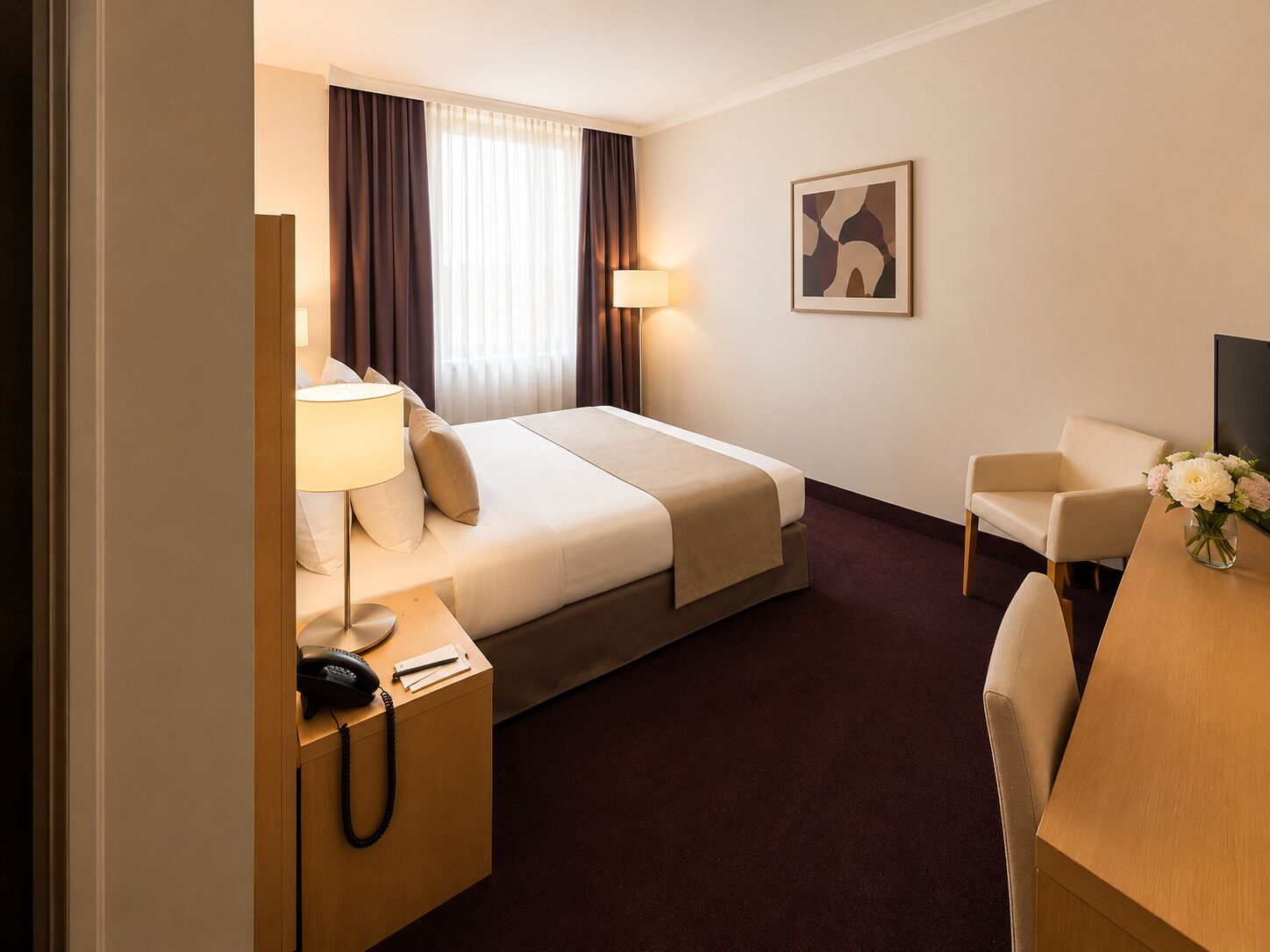 A hotel room with a bed, bedside table, lamp, chair, desk, and a vase with flowers.