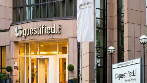 A hotel entrance with a large glass facade, a sign reading 'Guestified,' and a flag with the same logo. - guestified. GRAND AIRPORT Hotel (Ratingen)