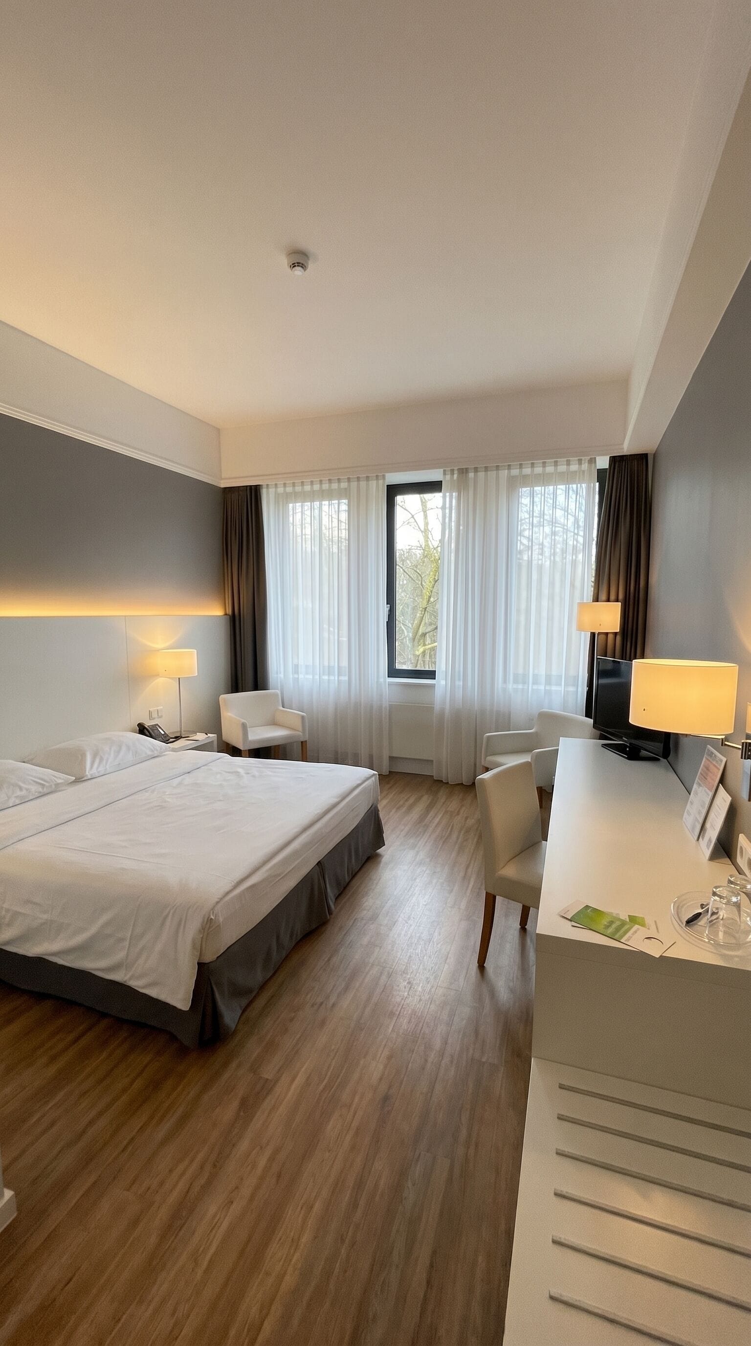 A hotel room with a large bed, two bedside tables, a desk with a chair, a television, and a window with curtains.