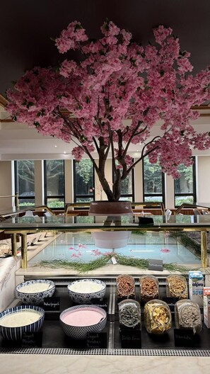 A restaurant with a large artificial cherry blossom tree, a glass display case with various food items, and a seating area with tables and chairs. - guestified. GRAND AIRPORT Hotel (Ratingen)