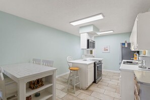 Una cocina compacta con armarios blancos, microondas, horno y lavamanos. Hay una mesa de comedor con sillas y un taburete pequeño.