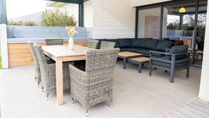 Une terrasse extérieure avec une table en bois, des chaises en osier, un canapé noir et un coin cuisine intégré.