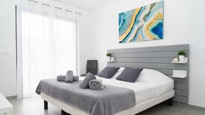 Une chambre moderne avec un lit, une literie grise, une tête de lit, une œuvre d’art accrochée au mur et une table de chevet agrémentée d’une plante.