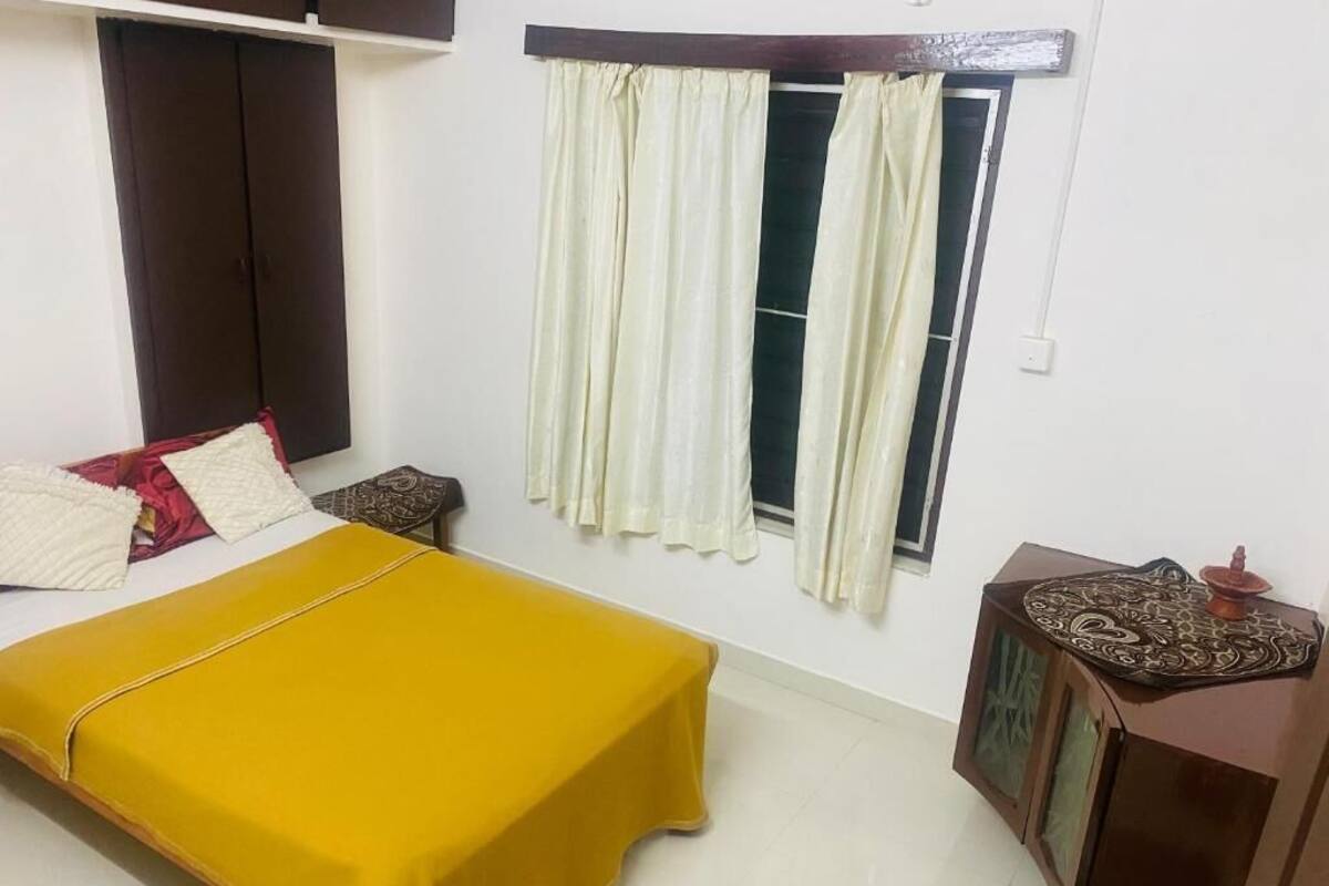 Un dormitorio con una cama, una mesita de noche, un armario, una ventana con cortinas y un estante en la pared.