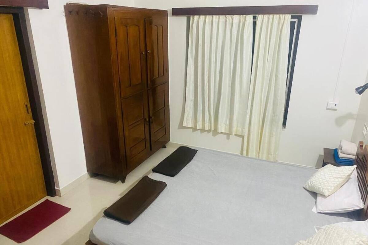 Un dormitorio con cama, armario, ventana con cortinas y mesita de noche con lámpara.