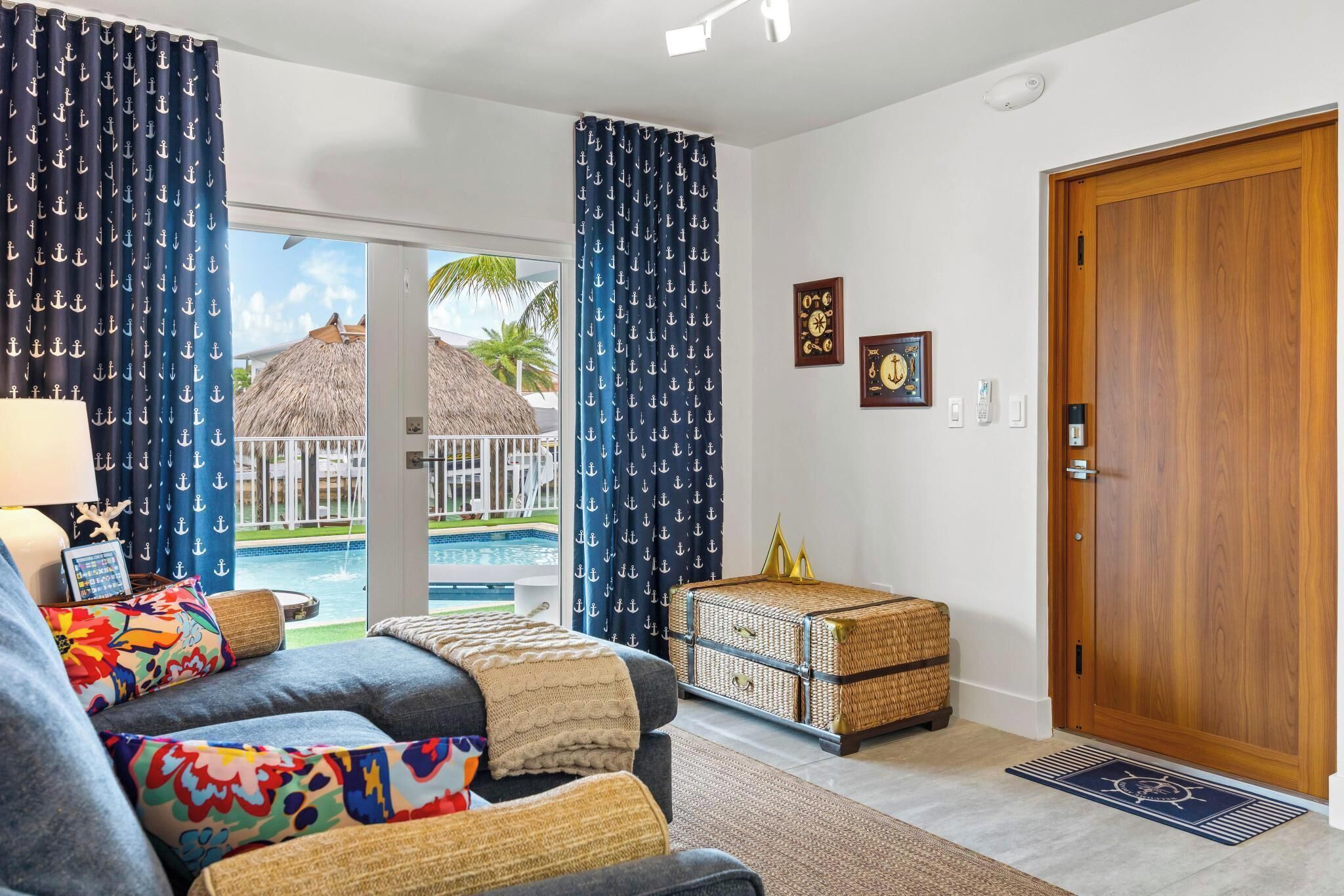 Ein Wohnzimmer mit einer Couch, Kissen, einer Korbo und einer Holztür, mit Blick auf einen Pool.