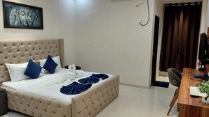 A hotel room with a tufted headboard, a bed with white linens, blue pillows, and a blue blanket. There is a desk with a computer and a small plant. - Hotel The White Palace (Lakhpat)