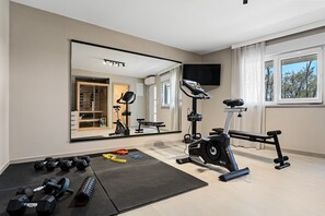 Ein Fitnessraum mit Trainingsgeräten, einem großen Spiegel und Blick auf Bäume durch das Fenster.