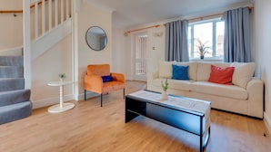 Un salon avec un escalier, un miroir rond, un canapé blanc agrémenté de coussins colorés, une table basse et une fenêtre avec des rideaux.