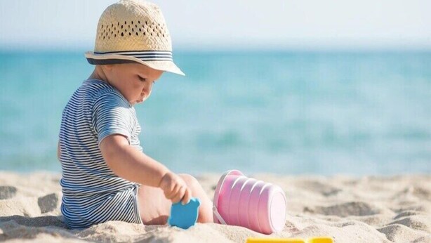 Ein Kind spielt mit Spielzeug an einem sandigen Strand, im Hintergrund ist ein klarer Blick auf den Ozean.