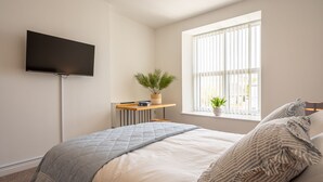 Ein Schlafzimmer mit einem Bett, einem an der Wand befestigten Fernseher, einem Fenster mit Jalousien, einem Nachttisch mit Pflanze und einer Nachttischlampe.
