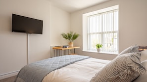 A bedroom with a bed, a TV mounted on the wall, a window with blinds, a bedside table with a plant, and a bedside lamp.