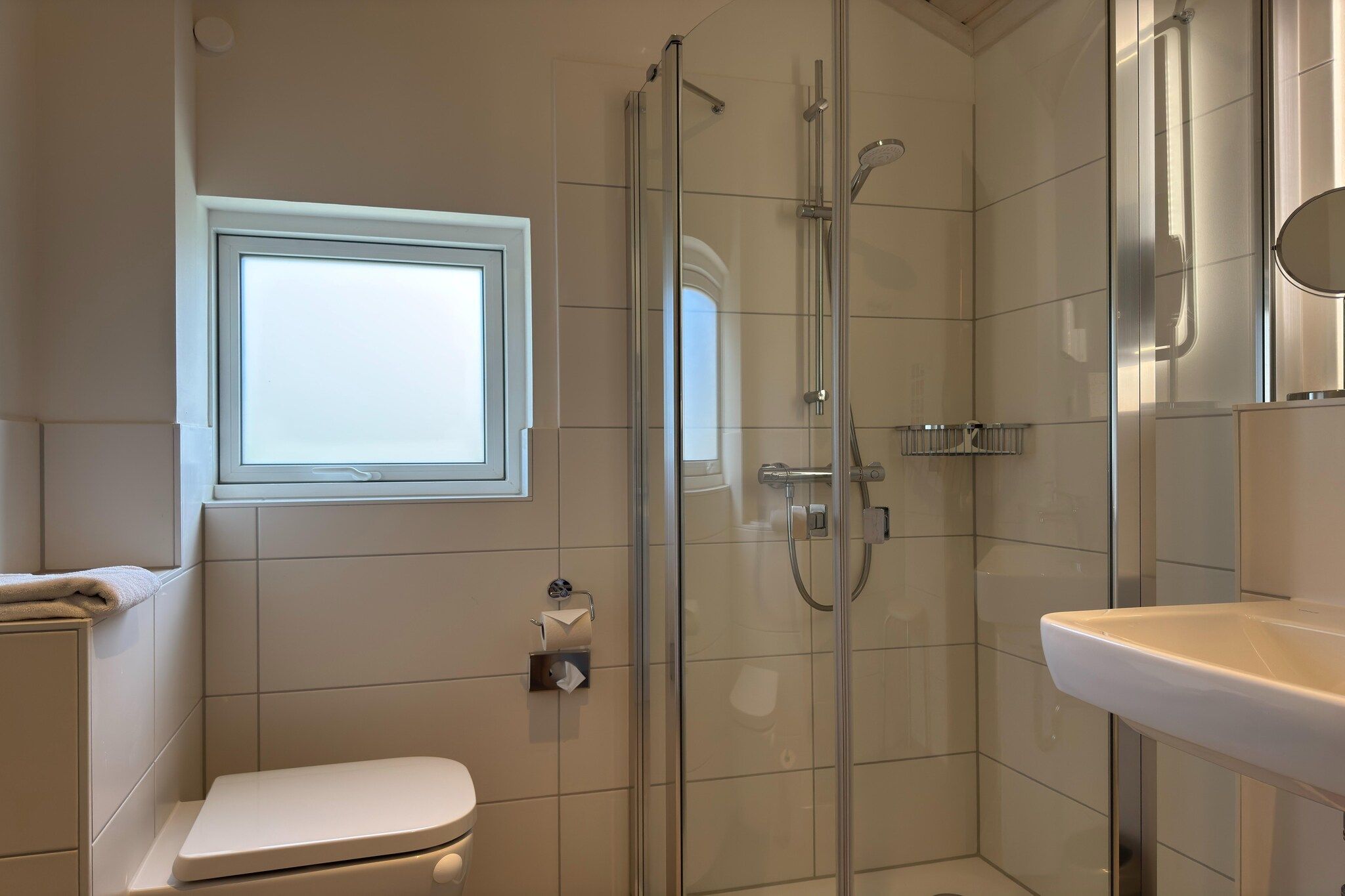 Un baño moderno con ducha envuelta en vidrio, inodoro, lavamanos y ventana que proporciona luz natural.