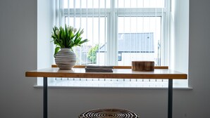 Een houten tafel met een witte vaas en een stapel papier, een houten kom, en een geweven mat voor een raam met jaloezieën.