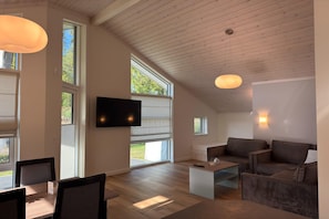Amplia sala de estar con televisor de pantalla plana, techo de madera, ventanas grandes y un acogedor rincón de descanso.
