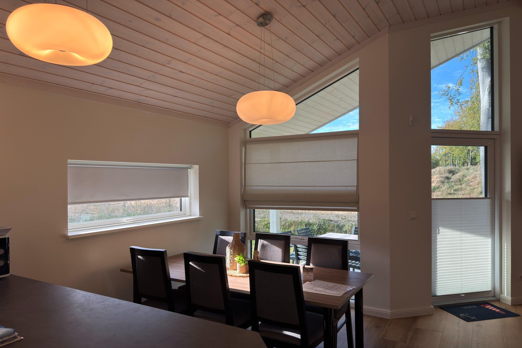 Ein Essbereich mit Tisch, Stühlen und Pendelleuchten. Es gibt Fenster mit Jalousien und Blick ins Freie.