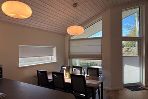 Un comedor con mesa, sillas e iluminación colgante. Se ven ventanas con persianas y vistas al exterior.