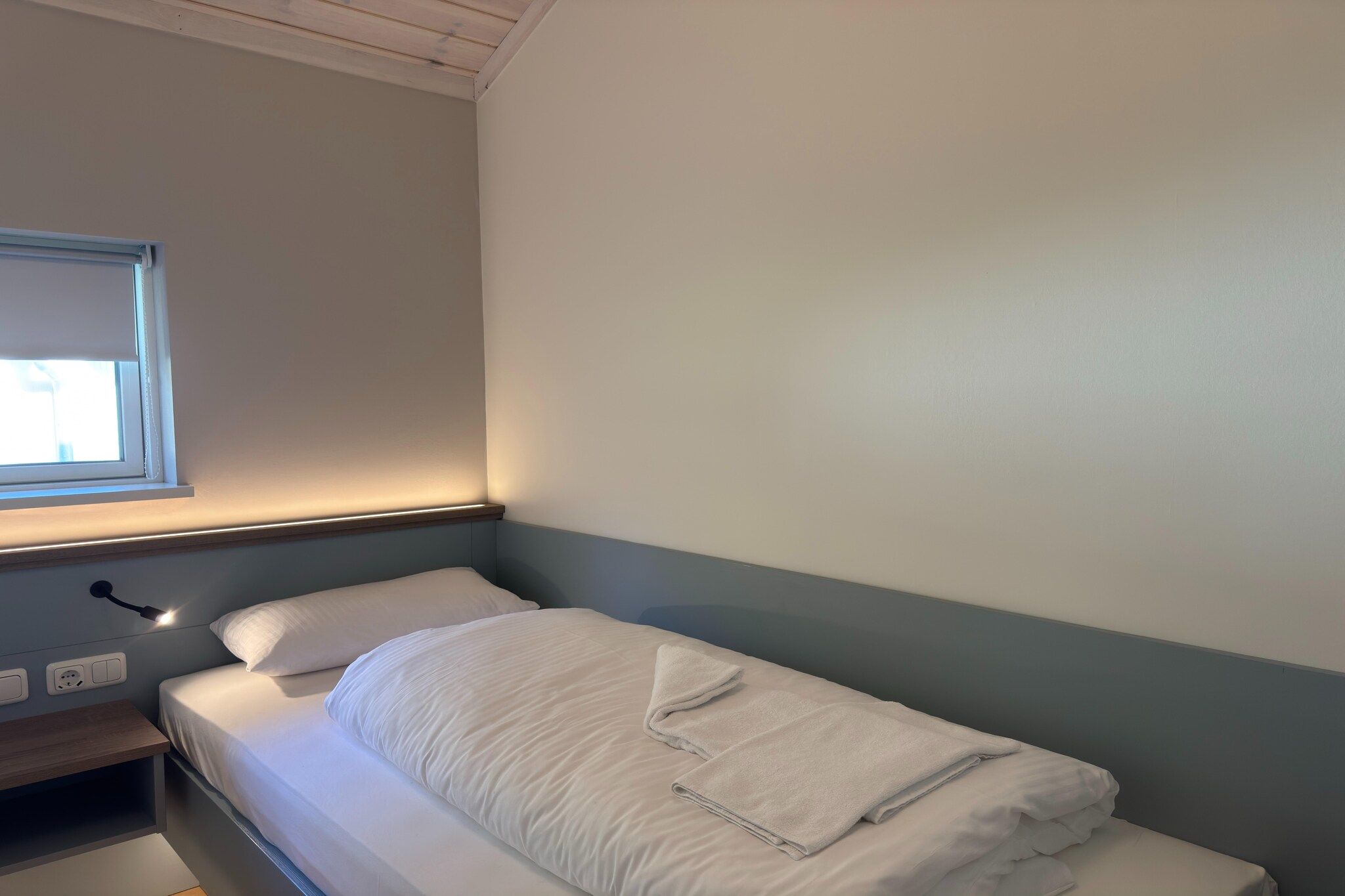 En säng med en vit madrass och kuddar, ett sängbord med en lampa och ett fönster med persienner.