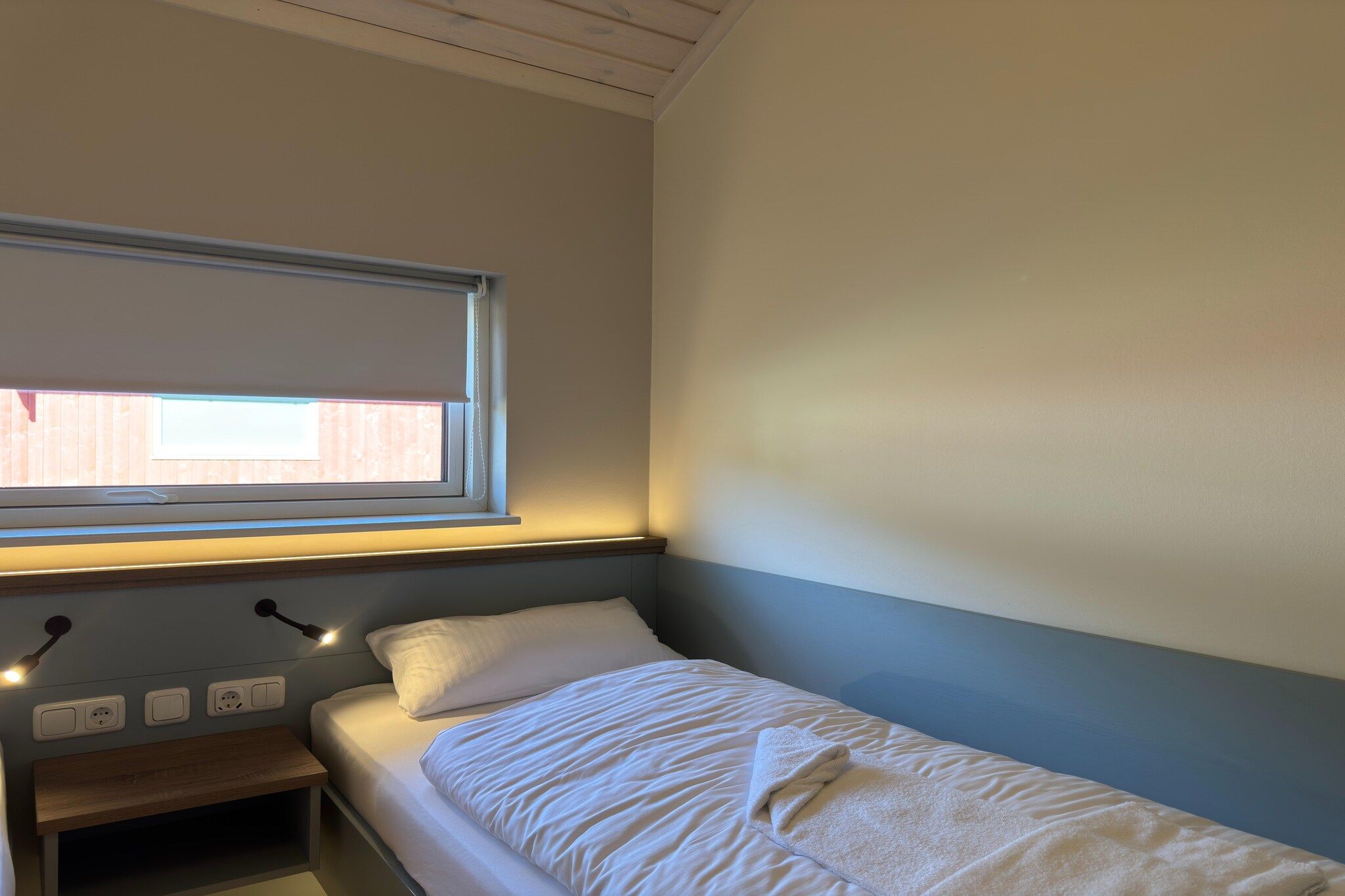 En enkelt seng med gavl, et natbord med lampe og et vindue med persienne.